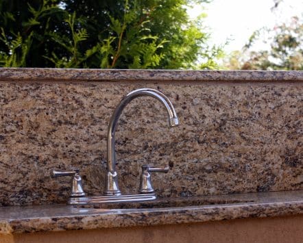 Chrome faucet plumbed into a granite outdoor kitchen countertop with greenery behind